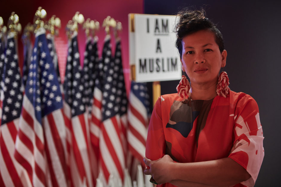 artist with arms crossed against a backdrop of US flags and a sign that says "I Am A Muslim"